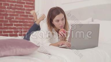 年轻的白人女人躺在床上，在<strong>笔记本</strong>电脑上打字，吃红<strong>苹果</strong>。 在家学习的漂亮女学生